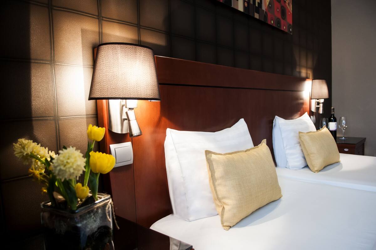 Quarto de hotel com cama bem arrumada, mesa de cabeceira com abajur e um vaso com flores.