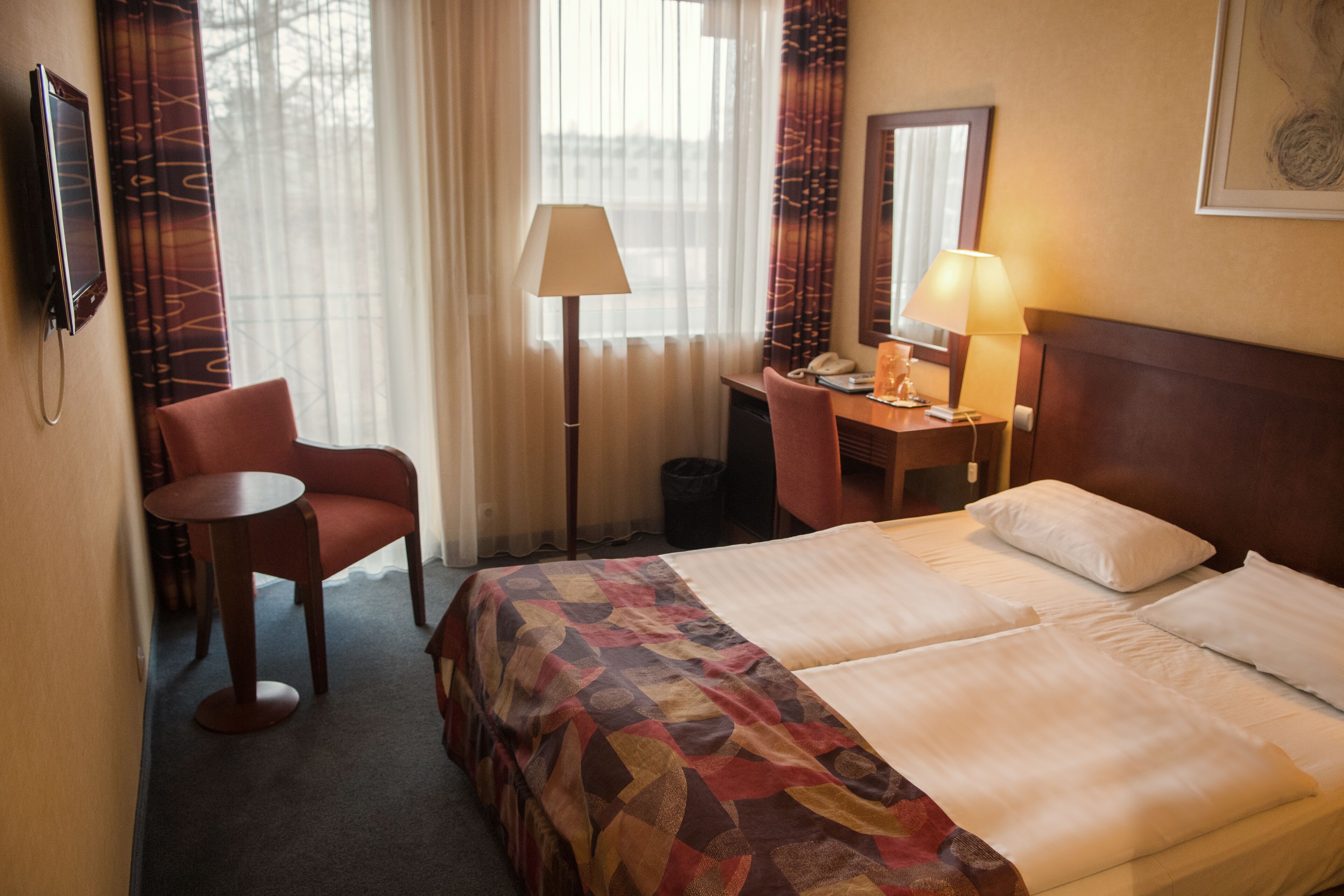 A hotel room with a bed, a desk, a chair, a lamp, and a TV.