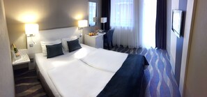 A hotel room with a large bed, bedside lamps, a desk with a chair, a TV mounted on the wall, and a window with curtains. - Hotel Azúr (Siófok)