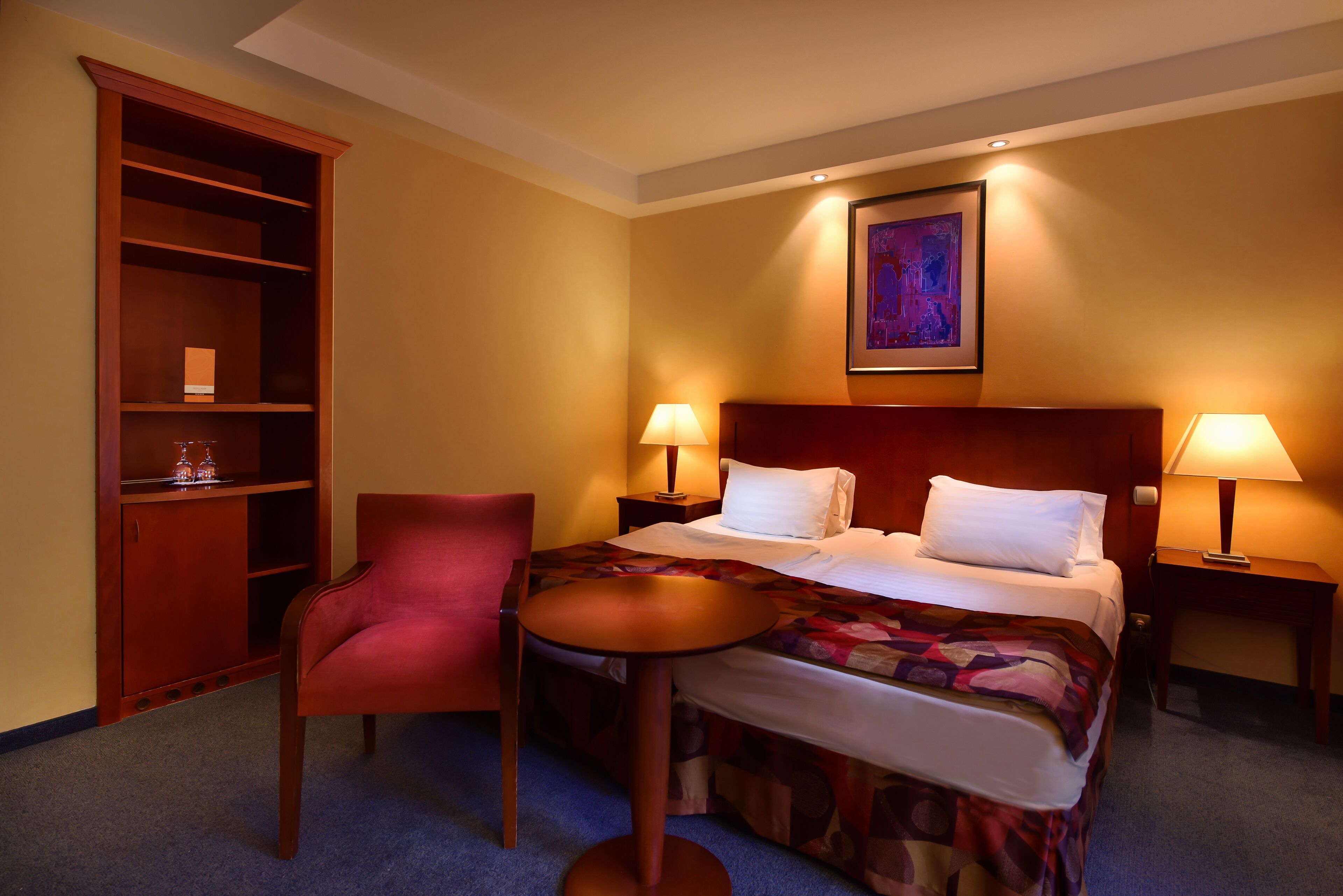 Habitación de hotel con una cama, dos lámparas de noche, una silla roja, un armario de madera y un cuadro en la pared.