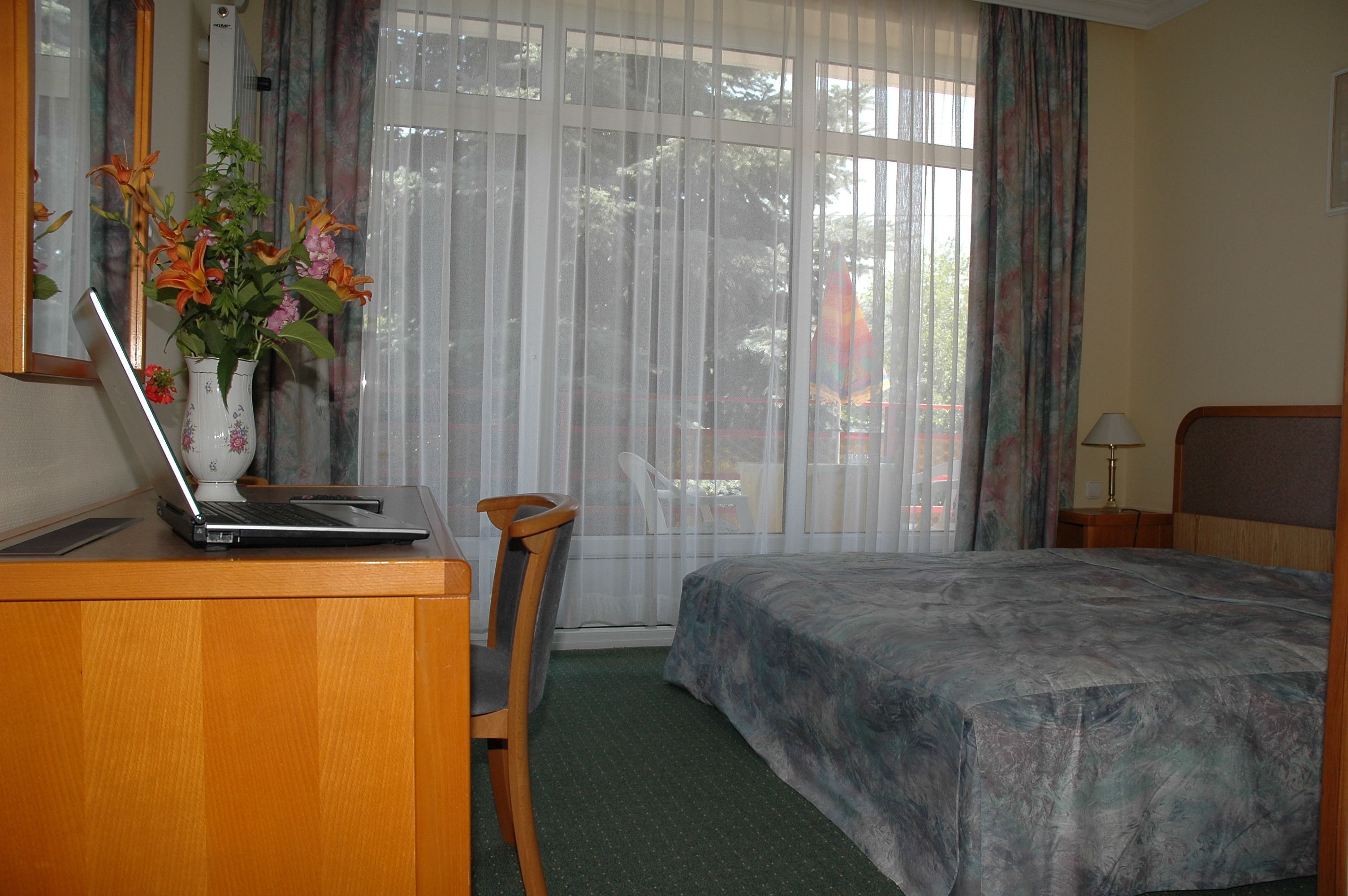 A hotel room with a bed, a desk with a laptop, a chair, and a vase with flowers.