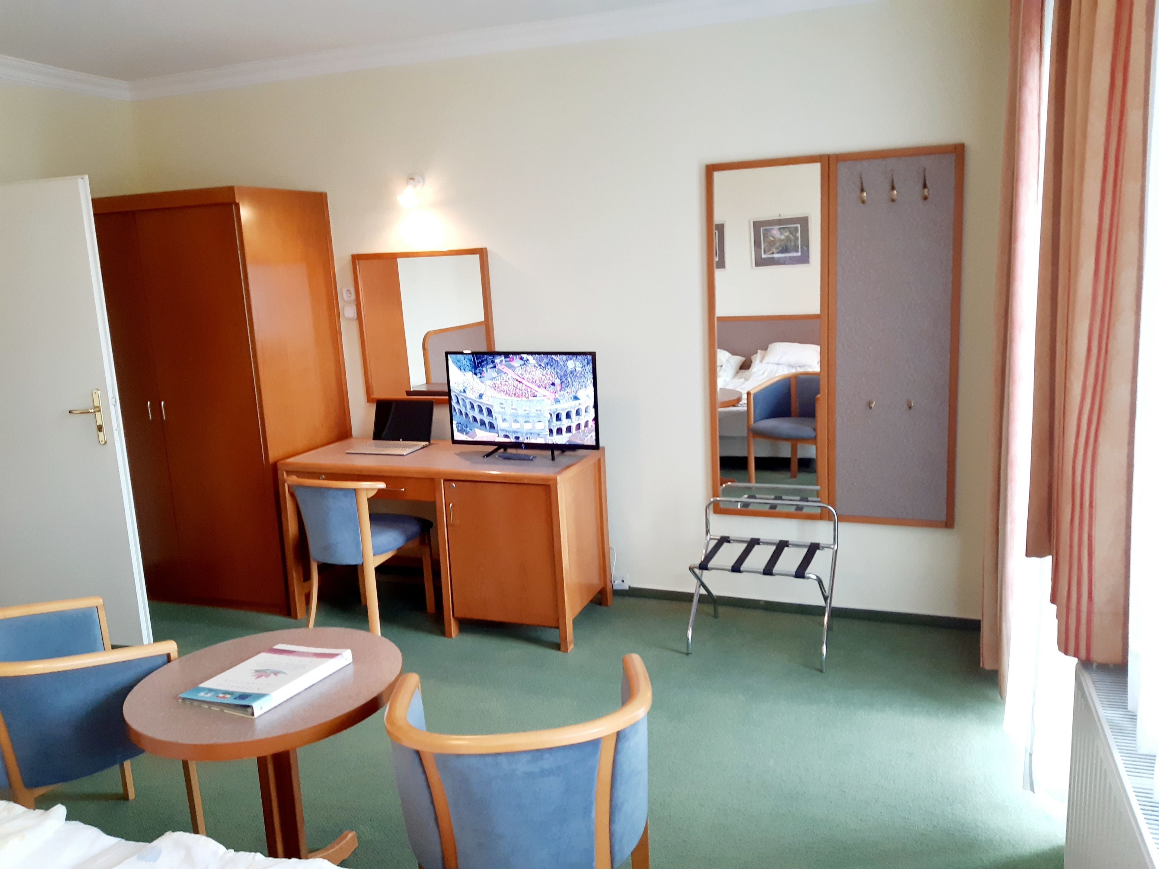 A hotel room with a desk, computer, chair, small table with a book, a mirror, and a wardrobe.