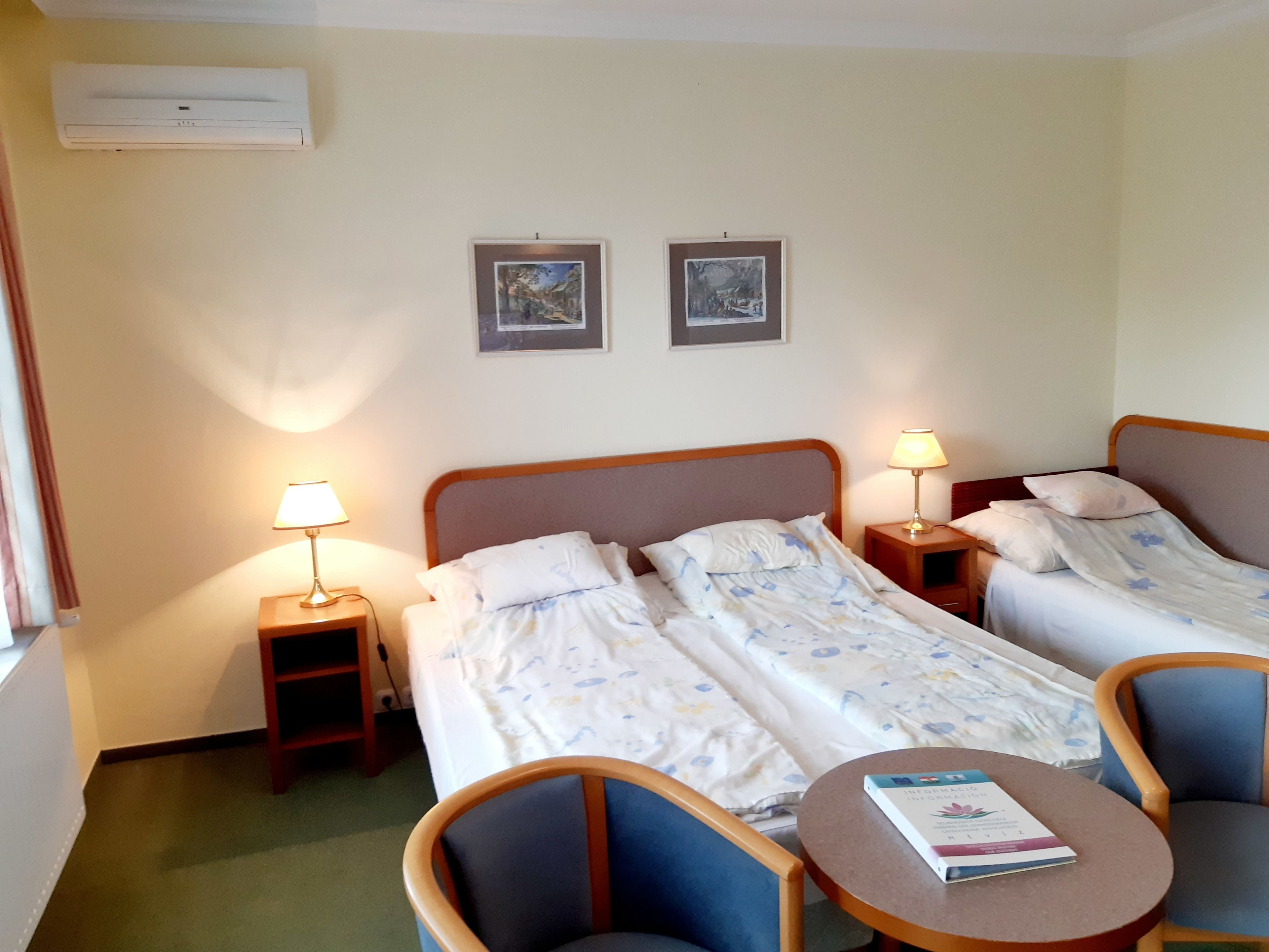 Una camera d'albergo con due letti, comodini con lampade, una sedia, un tavolino con un libro e due quadri appesi al muro.