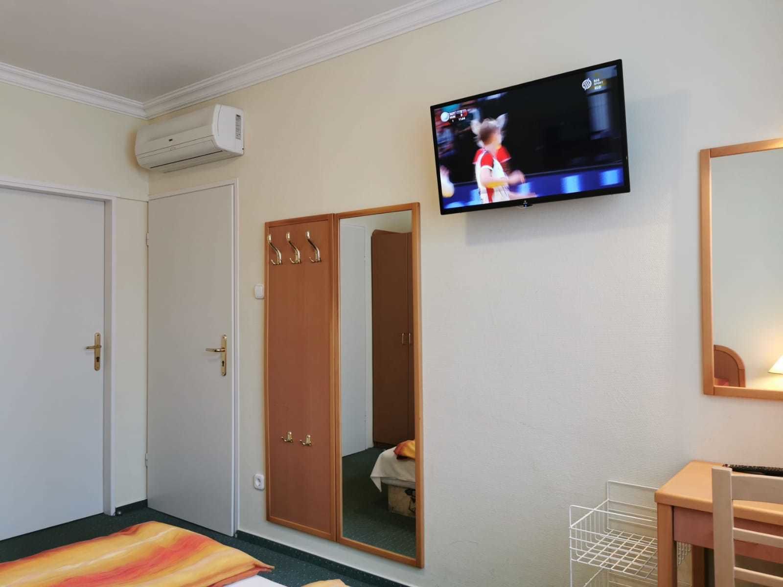 A hotel room with a bed, a TV mounted on the wall, a wardrobe with a mirror, and a desk with a chair.