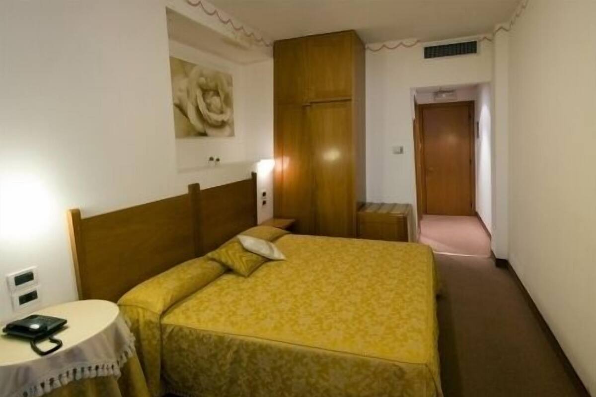 A hotel room with a bed, a bedside table, a lamp, and a door leading to another room.