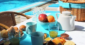 Un coin petit-déjeuner aménagé au bord d’une piscine, avec une chaise en osier, une table recouverte d’une nappe bleue, un pichet blanc, une tasse bleue, un verre de jus de fruits, des muffins et un saladier de fruits.