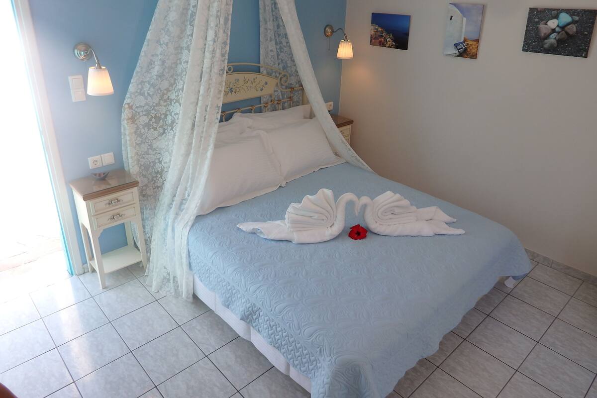 Uma cama bem arrumada com colcha azul e travesseiros brancos, uma mesa de cabeceira com abajur, e uma parede com quadros.