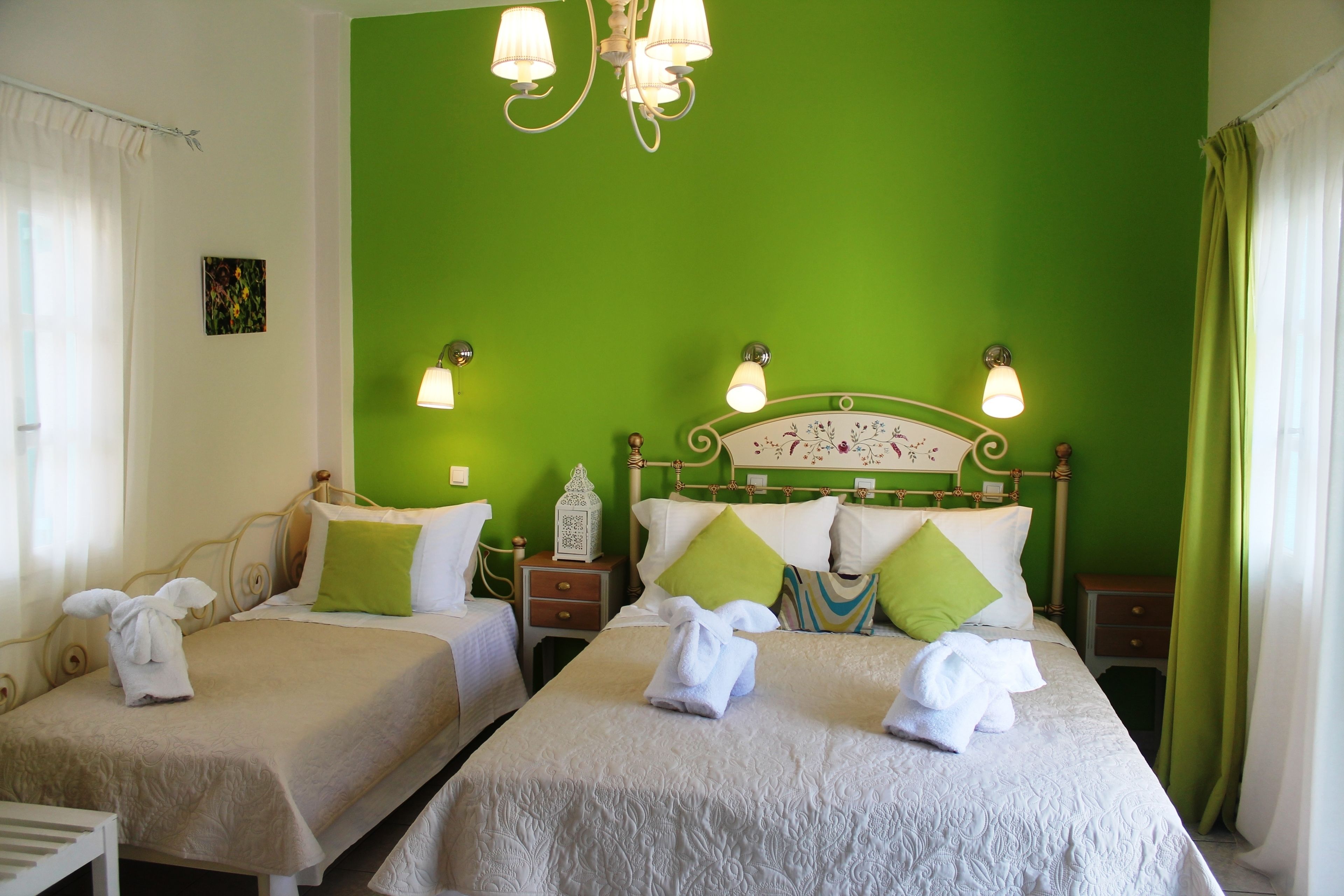 A hotel room with two single beds, each with a white bedspread and green pillows. There are bedside tables with lamps, a chandelier, and a small decorative item on one table.