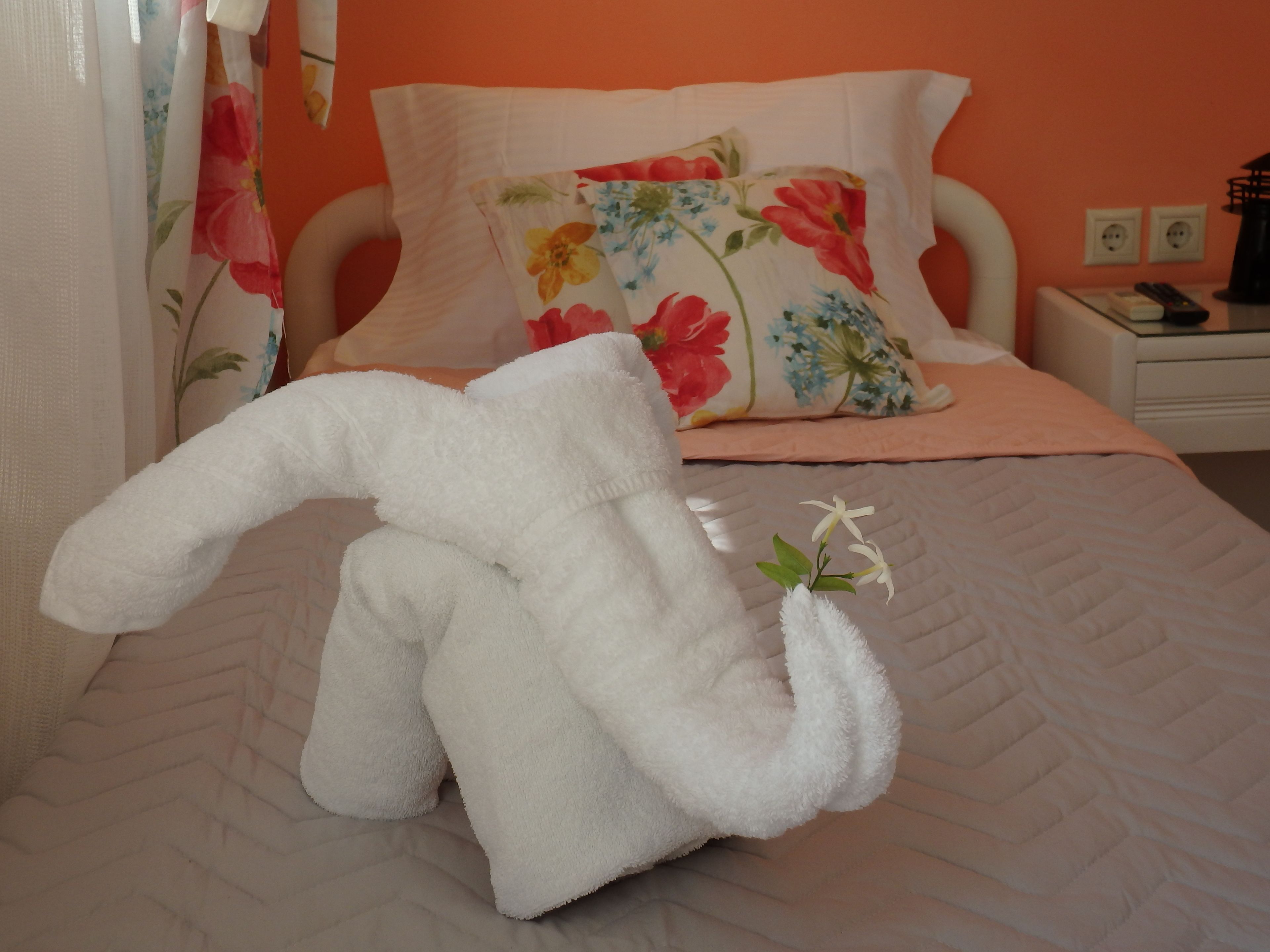 A bed with a floral pillow, a white fluffy towel, and a small white flower.