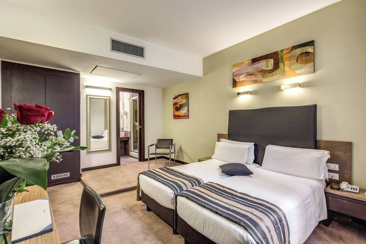 A hotel room with a large bed, bedside tables, a desk with a chair, a vase of flowers, and artwork on the walls.