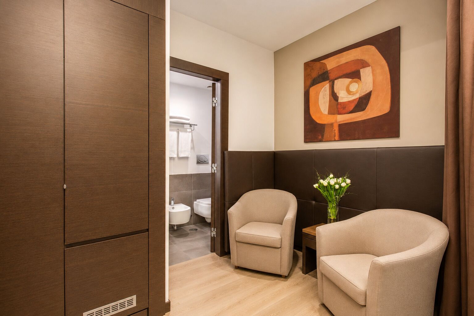 A hotel room with a wooden wardrobe, a seating area with two chairs, a vase with flowers, and a large abstract painting on the wall.
