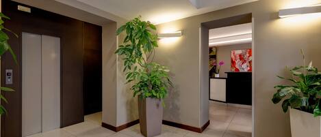 Um hall de hotel moderno com uma mesa de recepção, uma planta grande em vaso e um elevador.