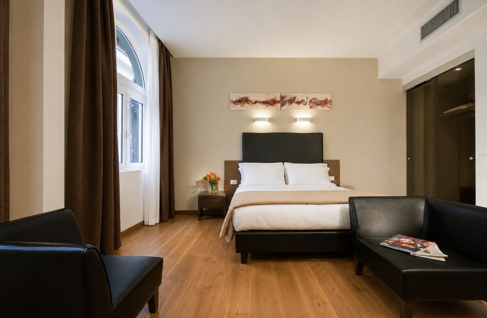 Une chambre d’hôtel avec un lit, deux chaises, une table de chevet et une fenêtre avec des rideaux.