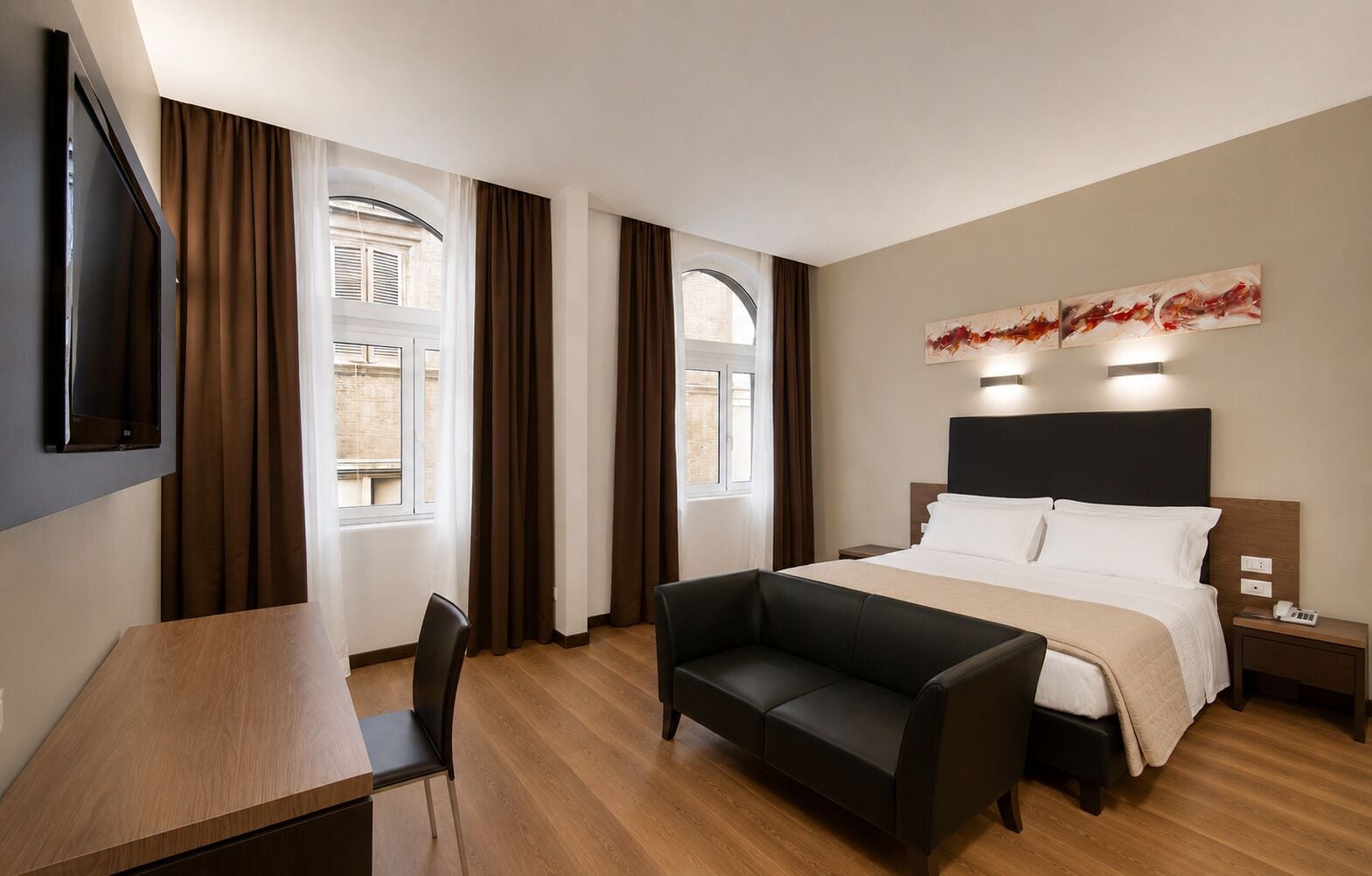A hotel room with a bed, a desk, a chair, a sofa, a TV mounted on the wall, and artwork above the bed.