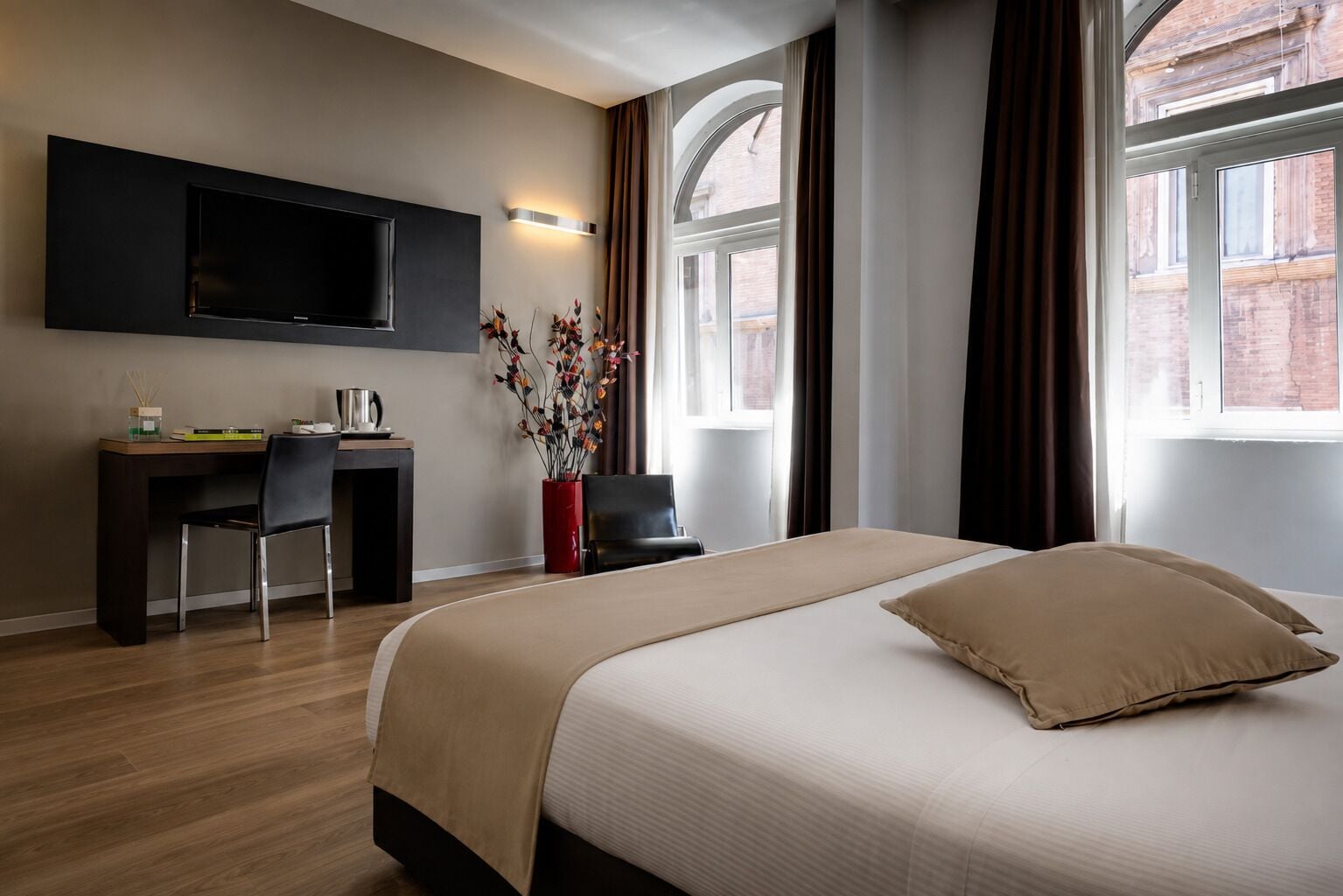 Une chambre d’hôtel moderne dotée d’un grand lit, d’un bureau, d’une chaise, d’un téléviseur à écran plat fixé au mur et d’une fenêtre avec des rideaux.