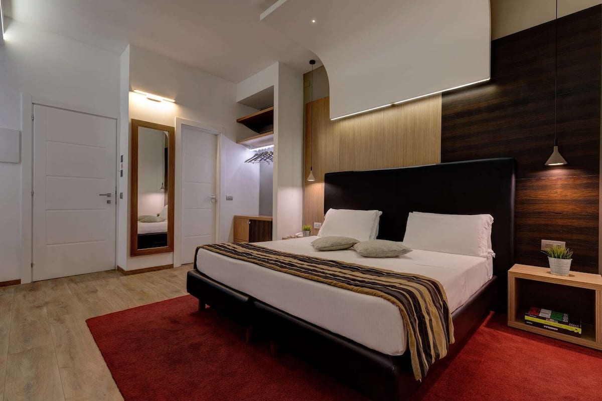 A modern bedroom with a large bed, a red carpet, a wooden headboard, and a small bedside table with a plant.