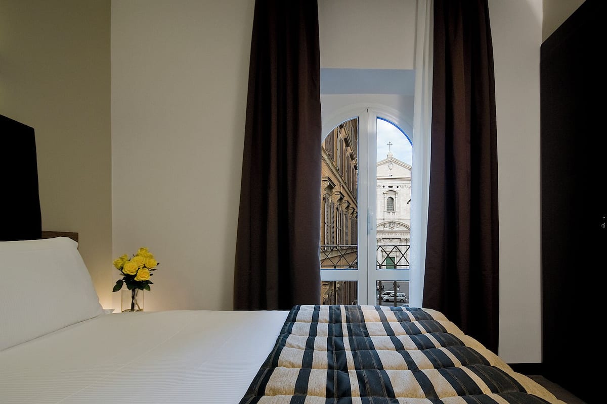 A hotel room with a bed, a vase of yellow flowers, and a view of a building through a window with curtains.