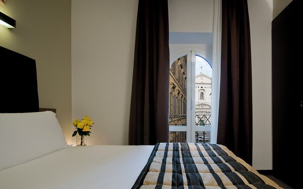 A hotel room with a bed, a vase of yellow flowers, and a view of a building through a window with curtains. - Hotel Rinascimento  (Rome)