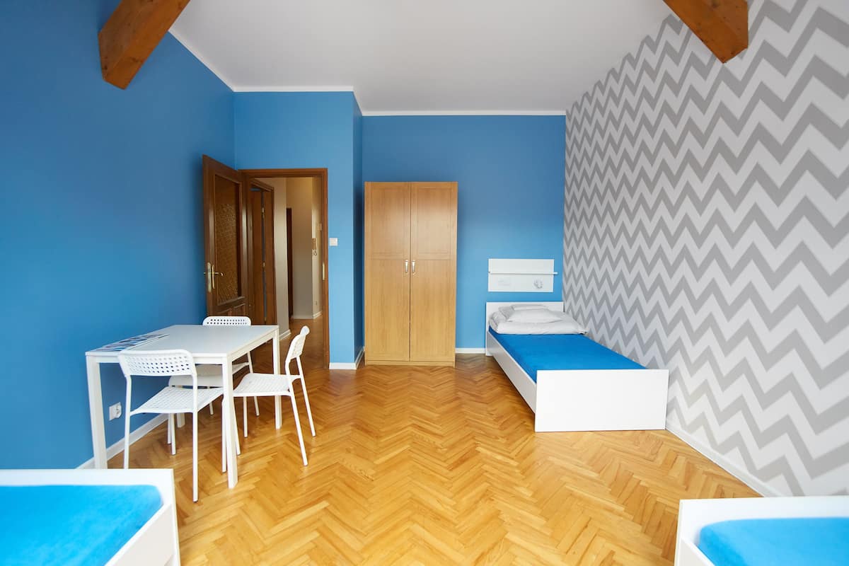 A room with a wooden herringbone floor, a white desk with chairs, a wardrobe, and a bed with a blue bedspread.