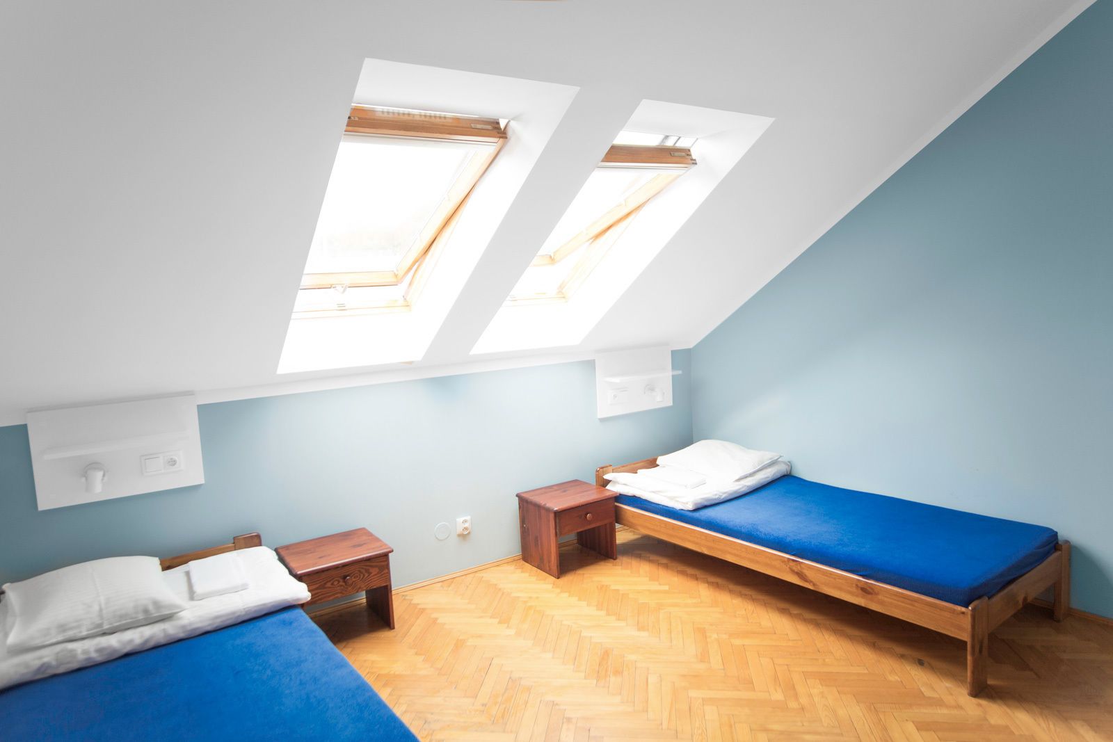 A dormitory room with two beds, a skylight, wooden flooring, and light blue walls.