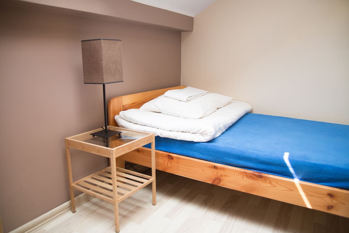 A single bed with a blue mattress and white bedding, a wooden bedside table with a lamp, and a wooden floor.