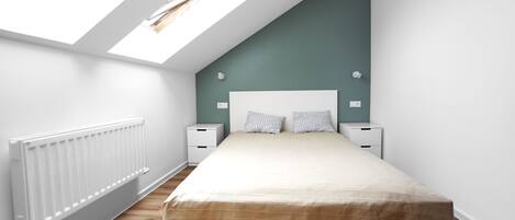 A bedroom with a sloped ceiling, skylights, a bed with a beige bedspread, two bedside tables with lamps, and a white radiator.