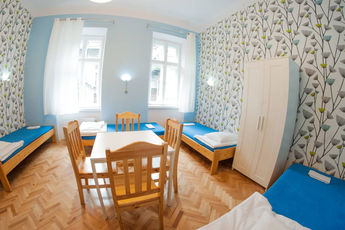 A room with two single beds, a wooden table with chairs, a wardrobe, and a window with curtains.