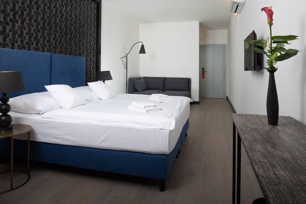 A modern hotel room with a large bed, a bedside table, a lamp, a small table, a mirror, and a vase with a flower.