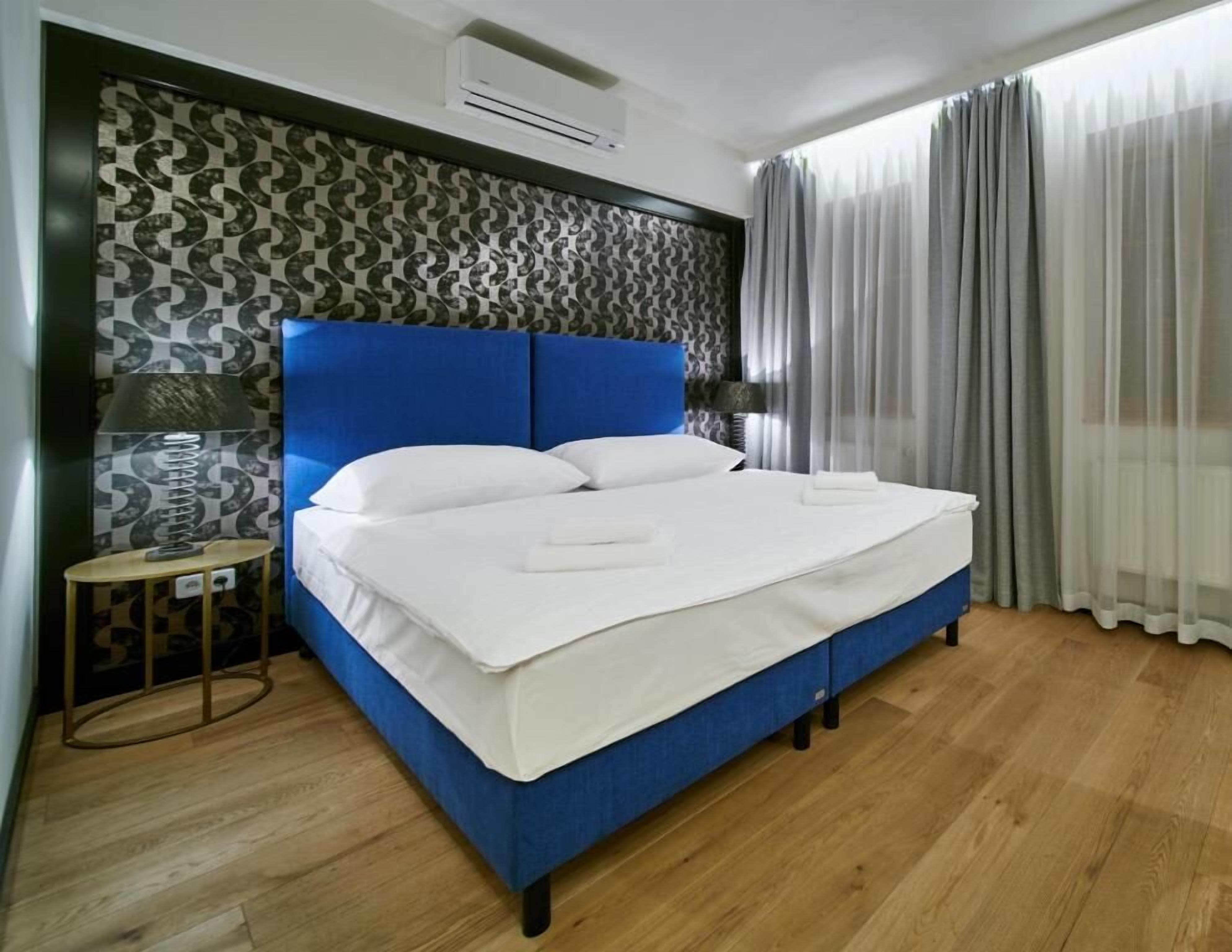 A neatly made bed with white linens, a blue headboard, and a patterned wall.