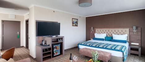 Habitación de hotel con cama, televisión, minibar, cafetera, frutero y mesa con una botella y vasos.