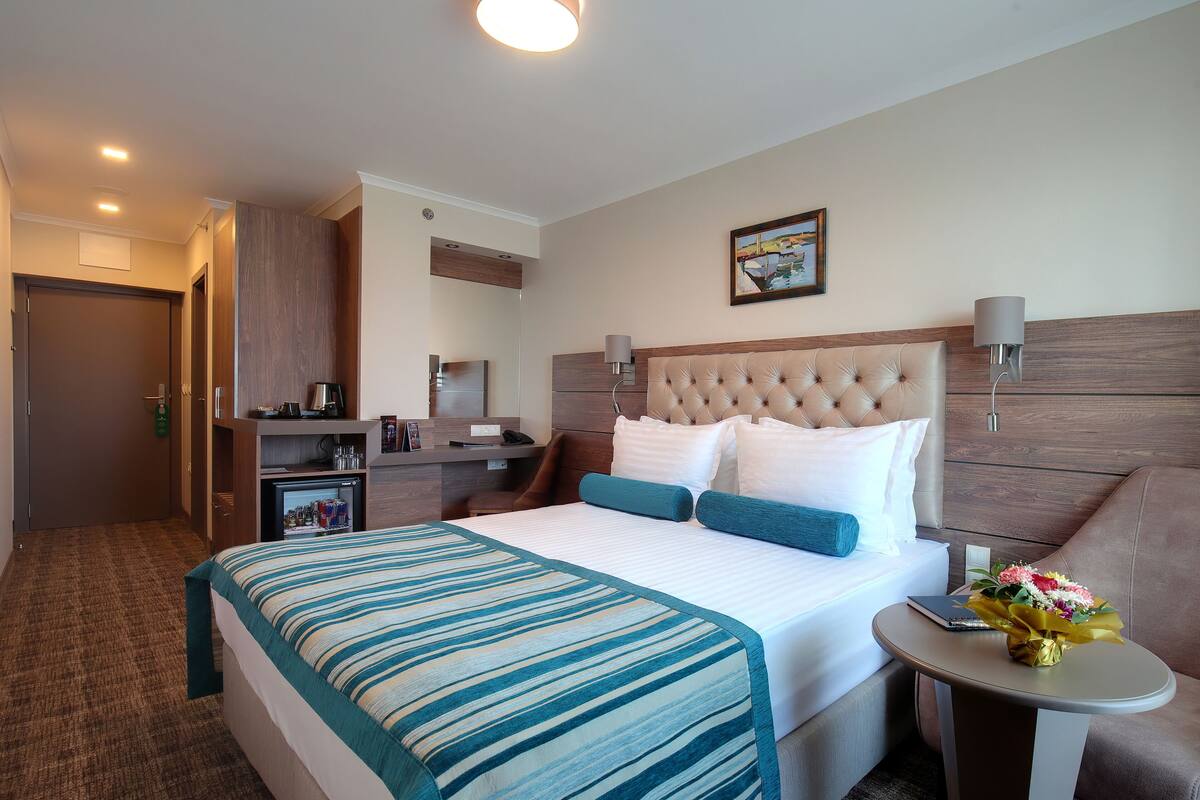 A hotel room with a large bed, a small desk, a chair, a bedside table with a lamp, and a wall-mounted TV.