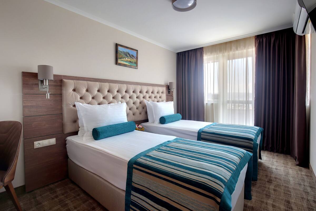 A hotel room with two beds, a headboard, a wall-mounted lamp, a painting, and a window with curtains.