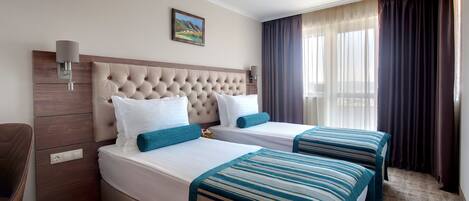 Habitación de hotel con dos camas, cabecero, lámpara de pared, un cuadro y una ventana con cortinas.
