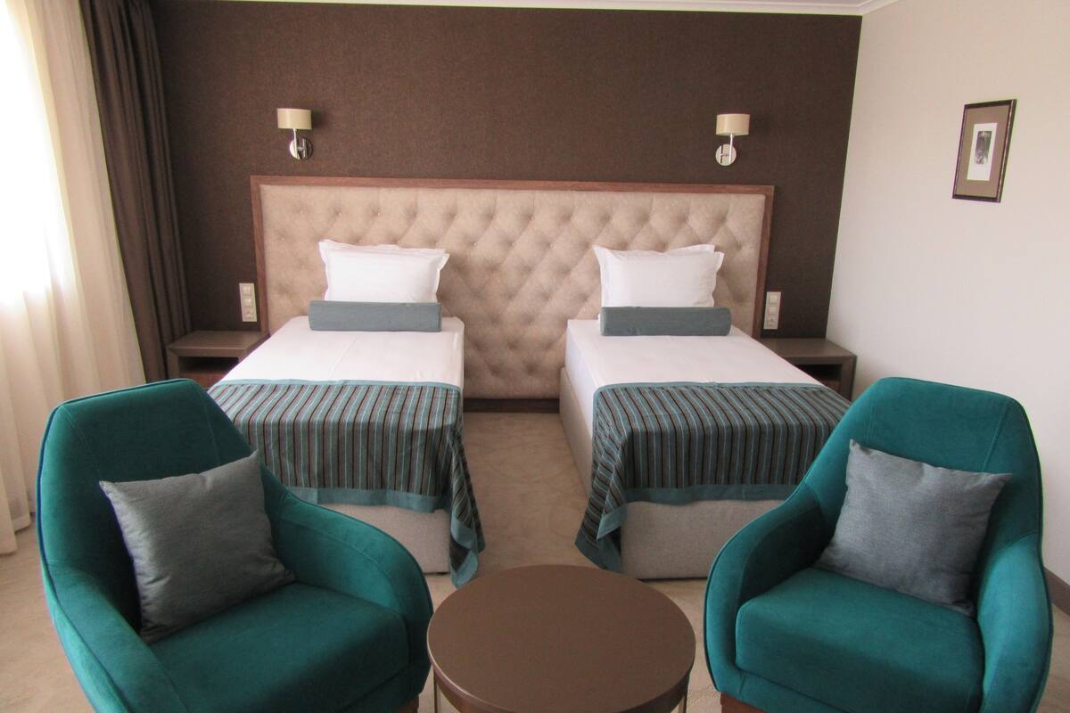 A hotel room with two beds, a round table, and two green armchairs.