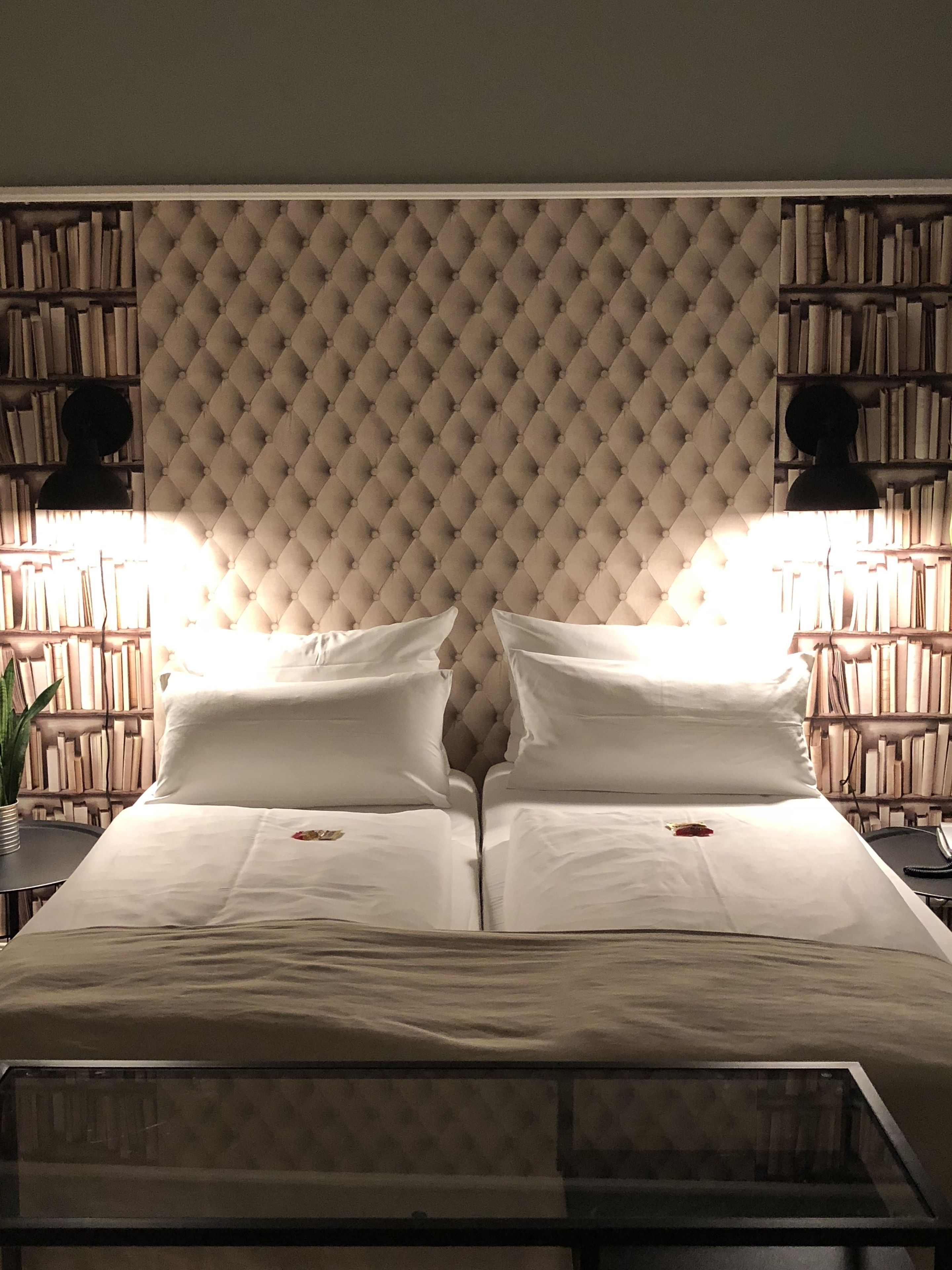 A double bed with a tufted headboard, flanked by two bookshelves filled with books, and a bedside table with a lamp.