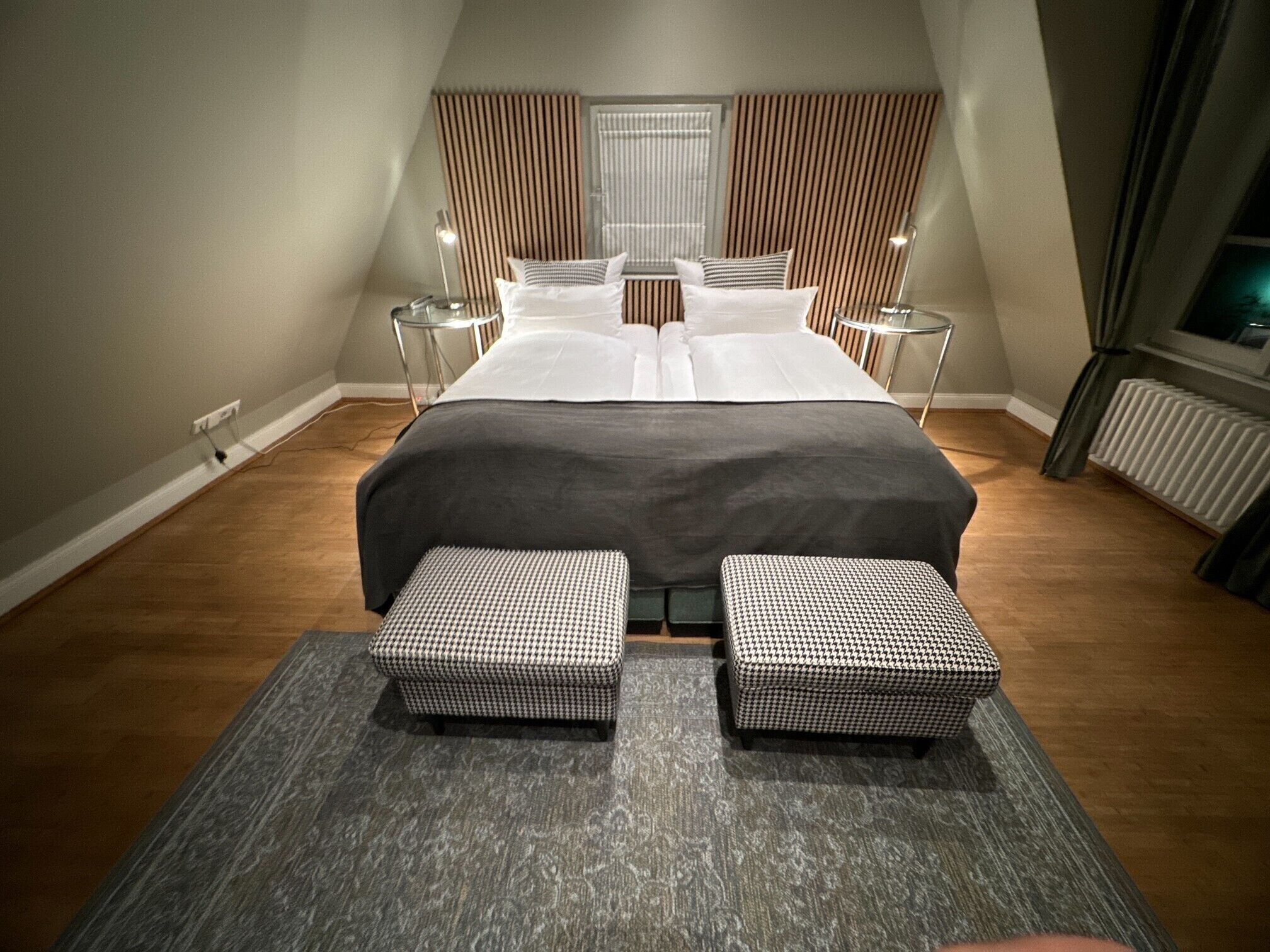 A modern bedroom with a large bed, two bedside tables, a window with blinds, and a radiator.