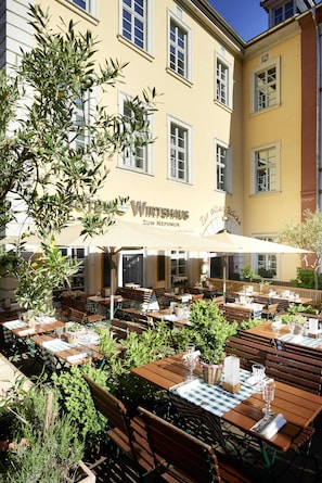 An outdoor cafe with wooden tables and chairs, surrounded by greenery, and a building with a yellow facade in the background. - Hotel Zur Alten Brücke (Heidelberg)