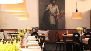 Interior de un restaurante con varias mesas, sillas, lámparas colgantes y un cuadro grande en la pared.