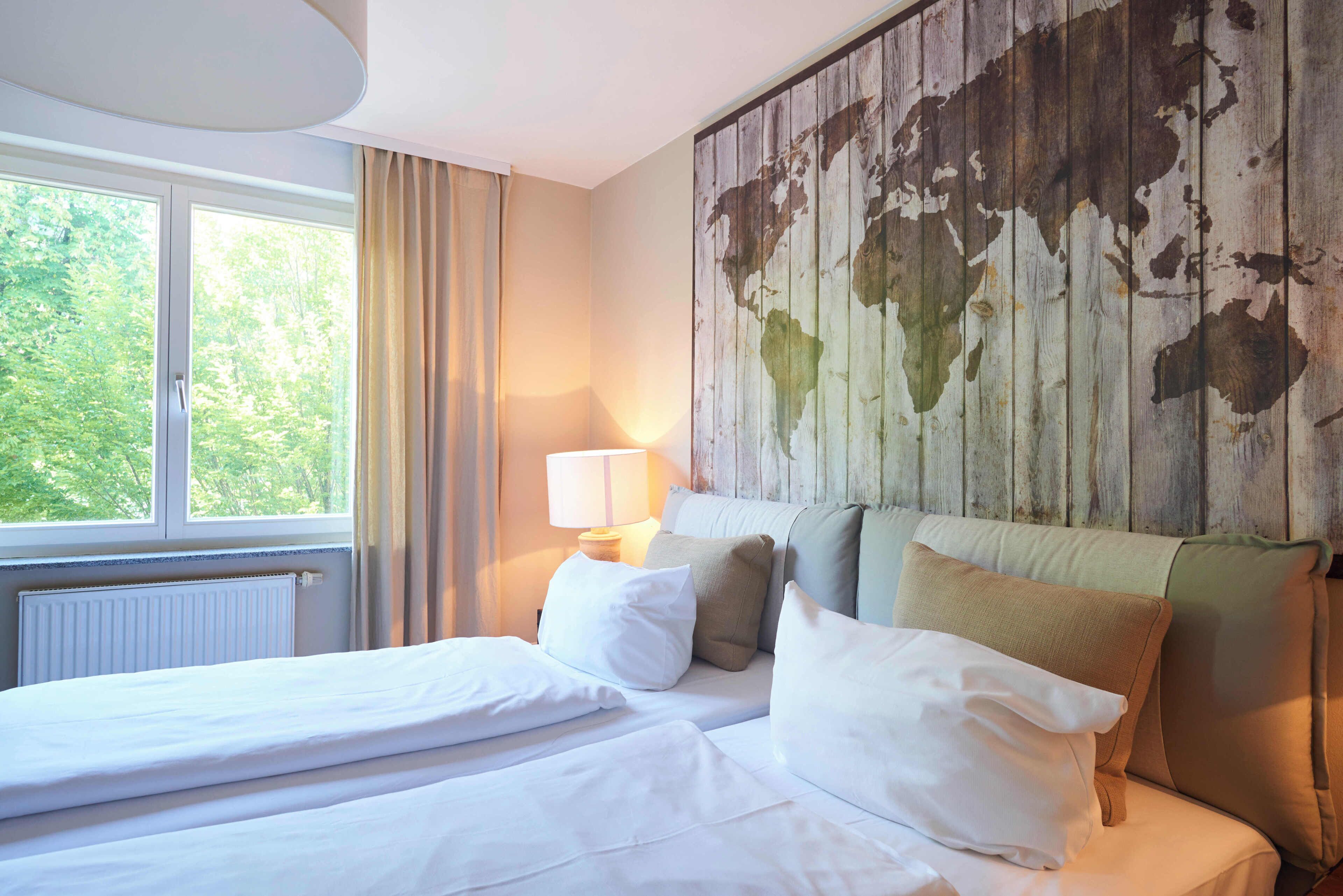 A hotel room with a bed, a window with curtains, a lamp, and a world map on the wall.