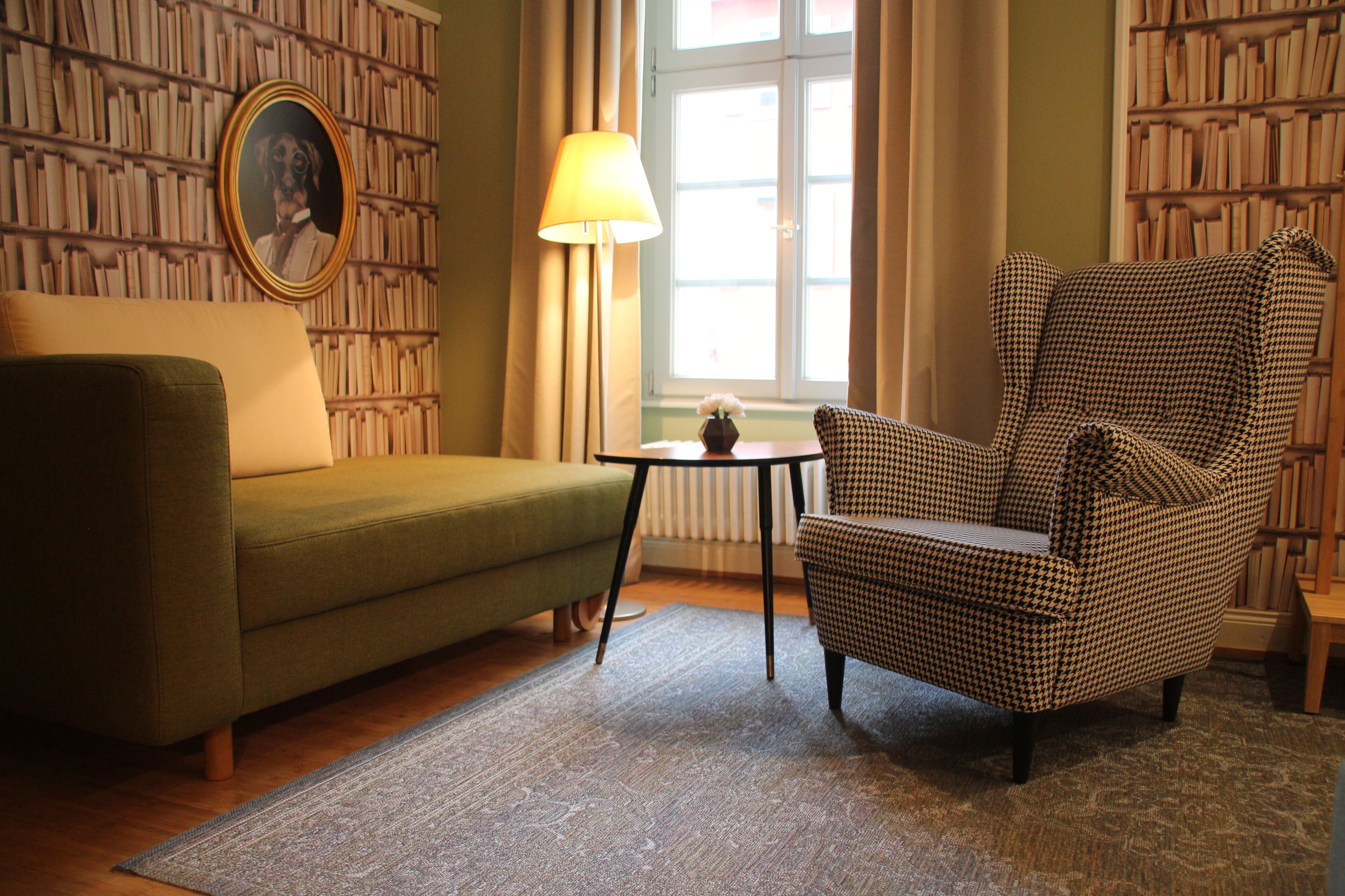 Ein Zimmer mit einem grünen Sofa, einem Hundszahn-Ohrensessel, einem kleinen Tisch mit einer Lampe und einem Bücherregal voller Bücher.