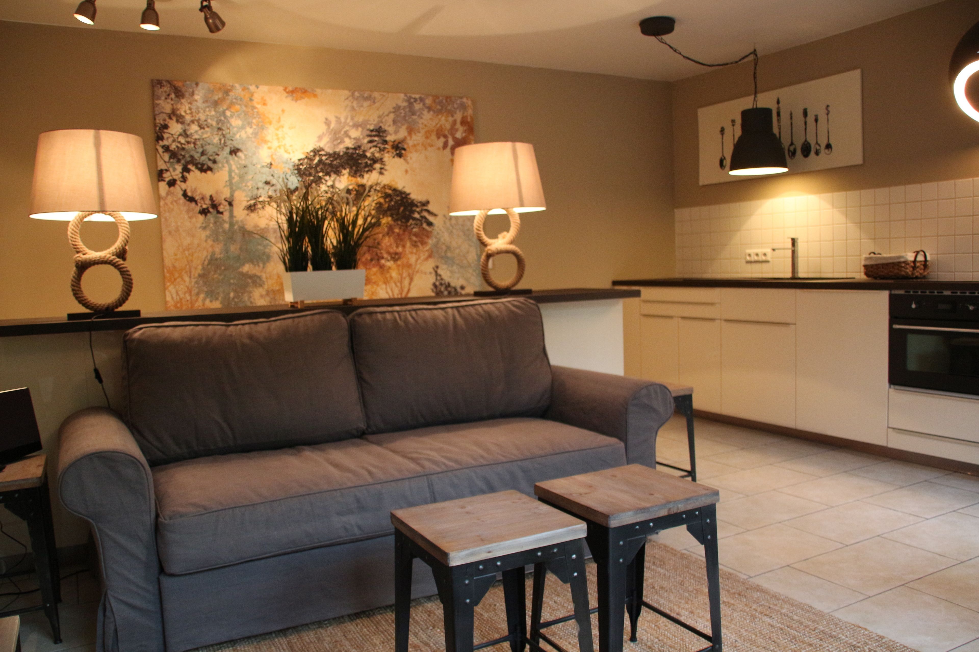 A living room with a sofa, two side tables, a painting, and a kitchen area with cabinets and a hanging light fixture.