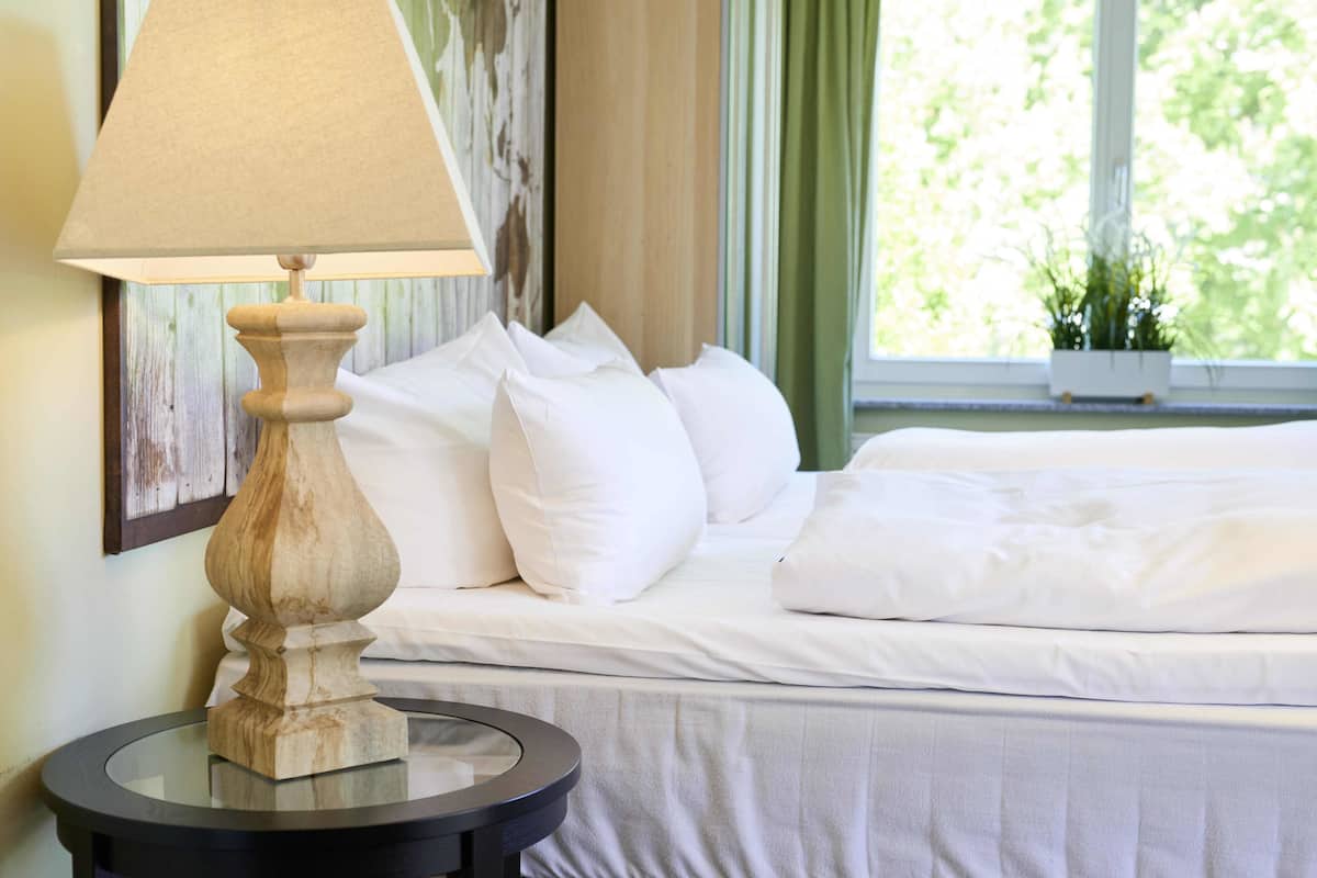 A neatly made bed with white linens and pillows, a round table with a lamp, and a window with curtains.