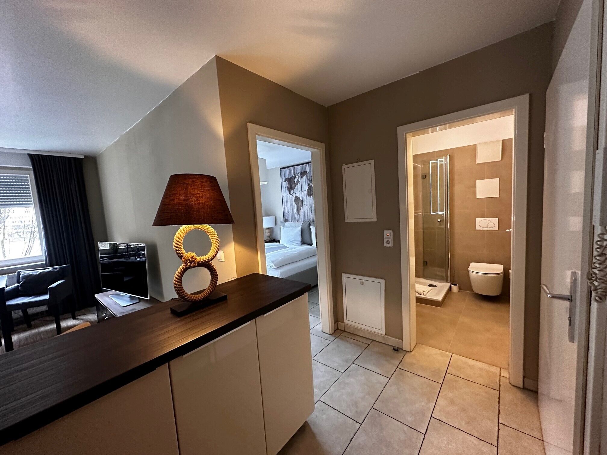 Habitació d'hotel moderna amb zona de cuina, taula de dinar, sofà, làmpada, dutxa i vàter.
