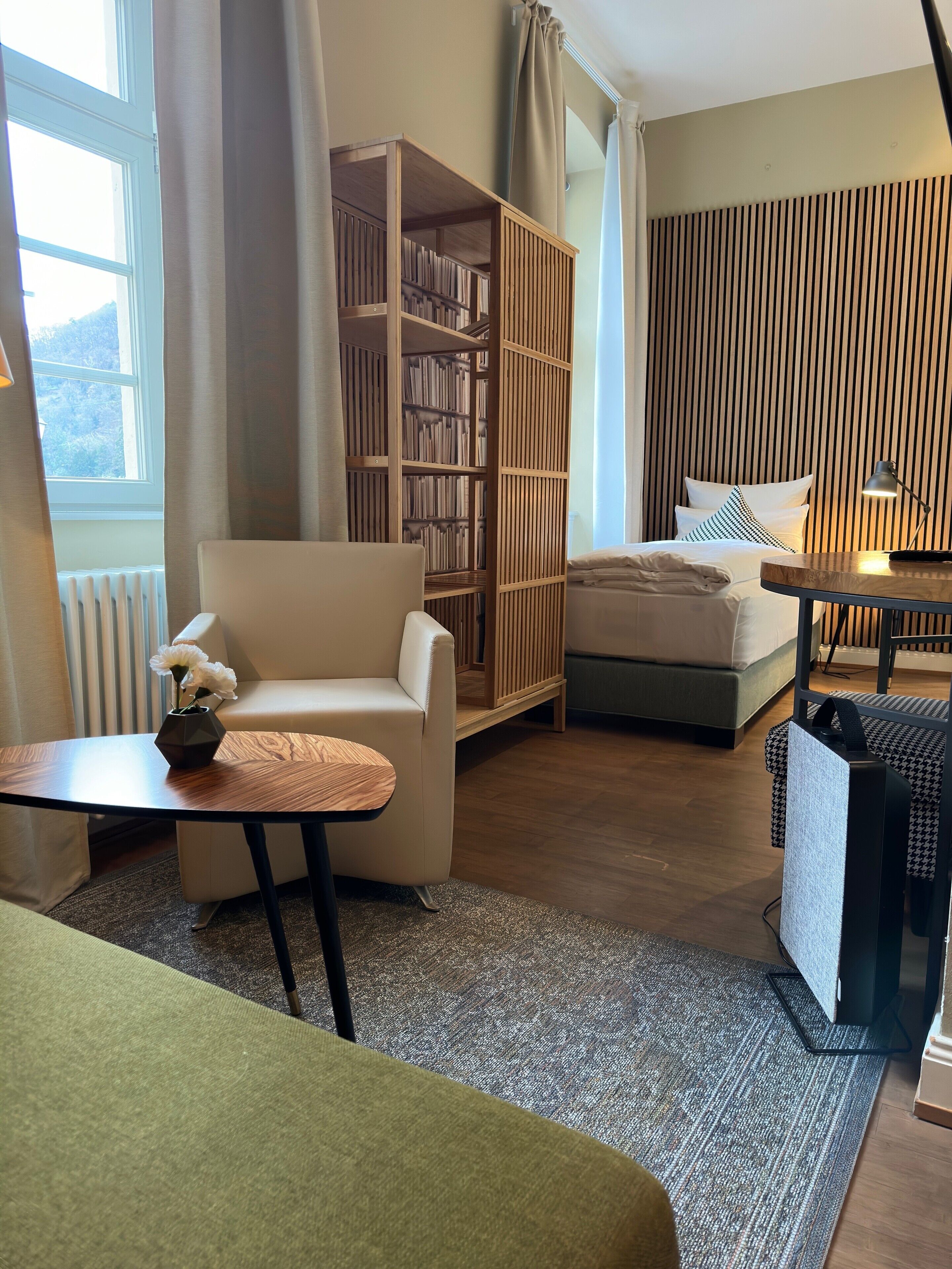 Ein modernes Hotelzimmer mit Bett, einer Sitzecke mit Tisch, einem Bücherregal und Blick ins Freie.