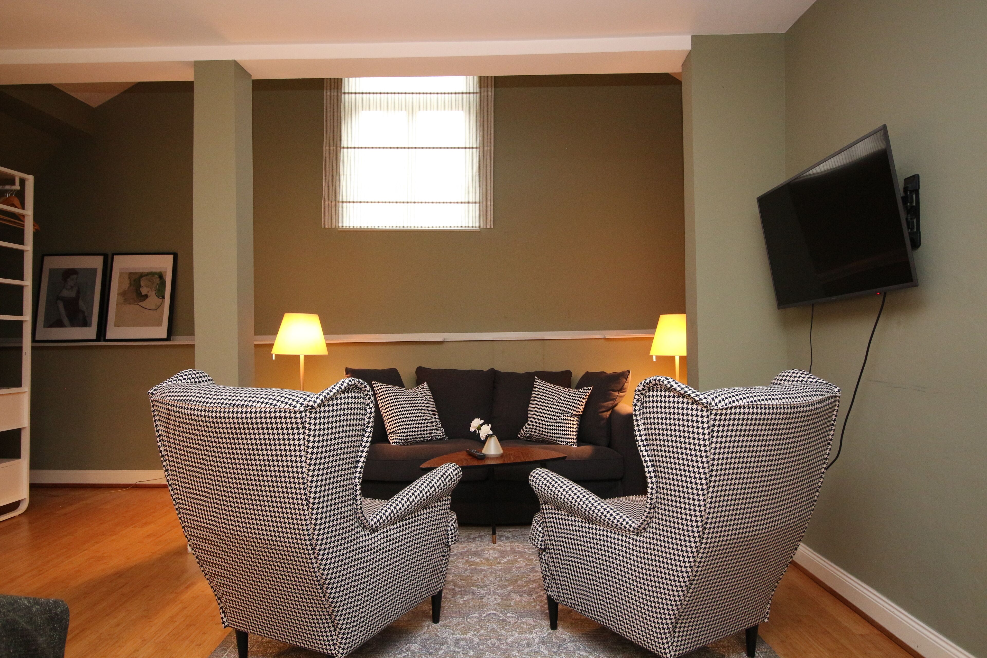 A living room with a sofa and two armchairs, a TV mounted on the wall, and a window with blinds.
