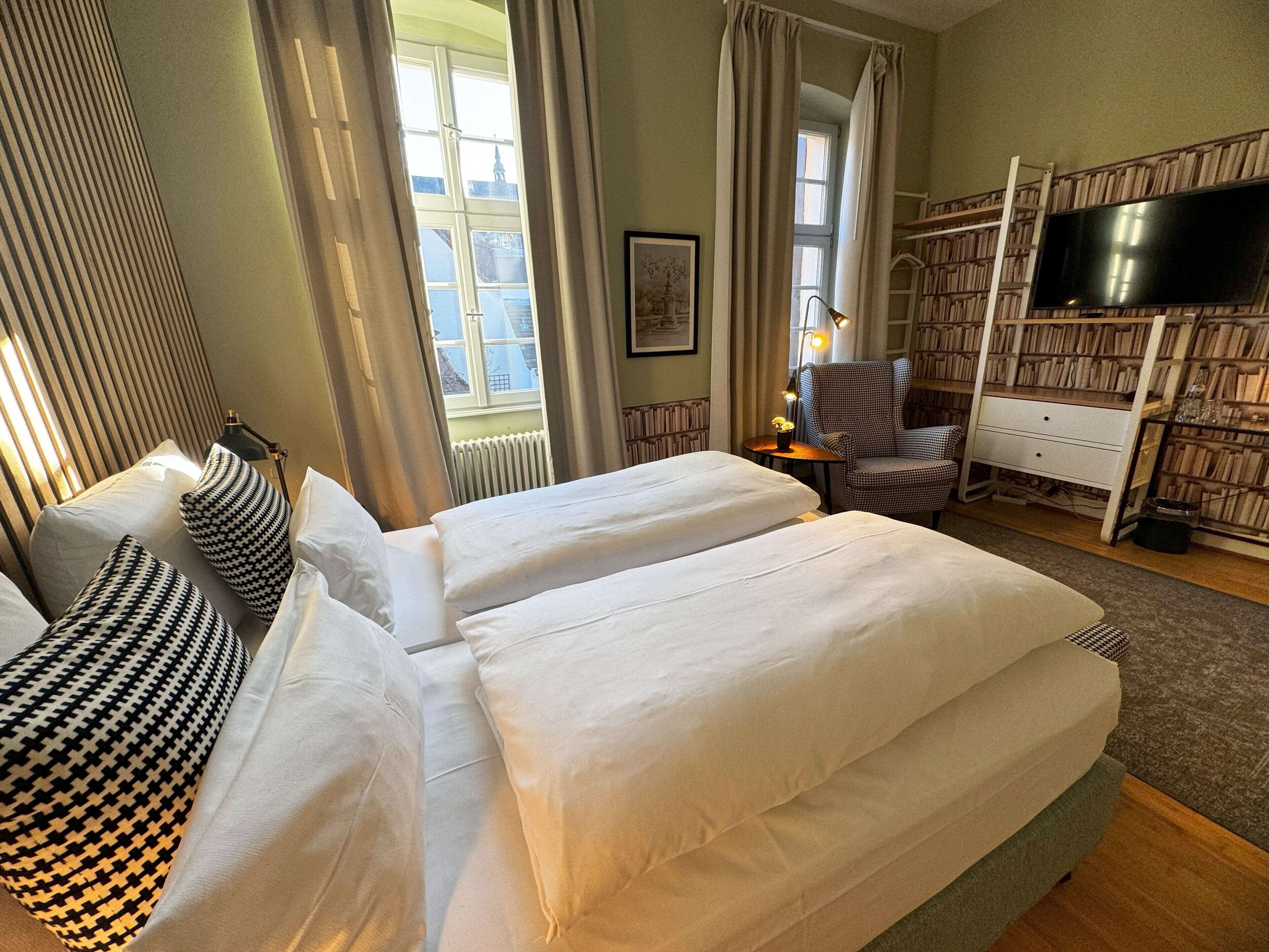 A hotel room with two beds, a chair, a desk with a TV, and a bookshelf.