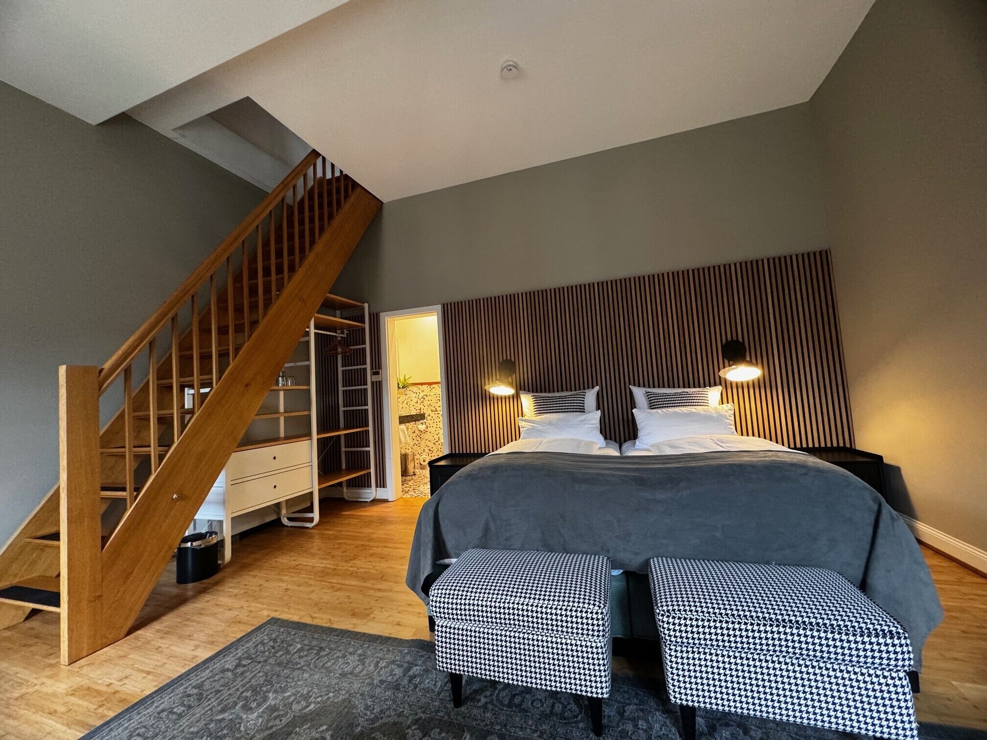 Ein Schlafzimmer mit einer Holztreppe, einem Bett mit weißer Bettwäsche, zwei Nachttischen und einem Kopfteil mit vertikalen Linien.