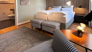 Un dormitorio moderno con cama, lámparas de noche, un sofá estampado con diseño pied-de-poule, una mesita con un jarrón y vistas al baño.
