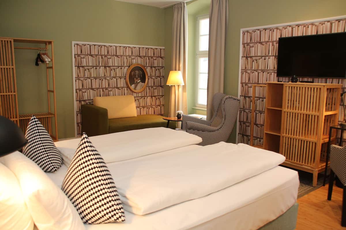 A hotel room with a large bed, a chair, a desk, a bookshelf, and a television.