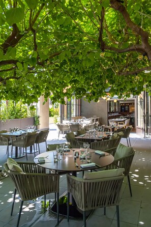 Un espace de restauration en plein air, avec des tables rondes et des chaises en osier, entouré d’une végétation luxuriante.